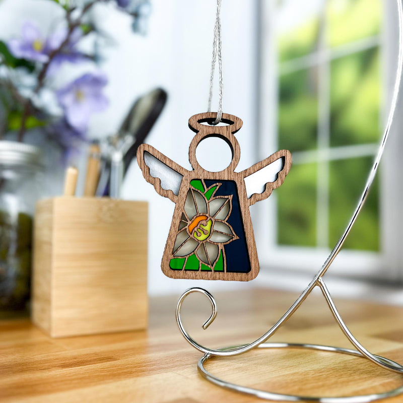 The Forged Flare® Narcissus Ornament, a 3.5" Mother's Angels® figurine with a stained-glass narcissus flower and "SV" initials, hangs from a silver stand on a wooden desk near a window with blurred flowers in the background.