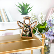 The Forged Flare® Narcissus Ornament—a 3.5" Mother's Angels® angel figurine—hangs on a gold shelf among books, a potted plant, and jars of flowers in a bright, modern room.
