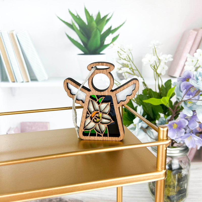 The Forged Flare® Narcissus Ornament—a 3.5" Mother's Angels® angel figurine—hangs on a gold shelf among books, a potted plant, and jars of flowers in a bright, modern room.
