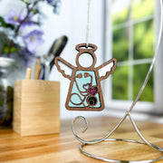 The Forged Flare® Mother's Angels® 3.5" wood angel ornament features a heart and paw cutout, hanging on a silver stand—perfect as a doctor or nurse gift—and is displayed on a wood surface with flowers, desk items, and a window in the softly blurred background.