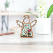 The Forged Flare® Mother's Angels® 3.5" Angel Figurine features a cutout stethoscope and pink glitter heart, making it a charming Doctor/Nurse Ornament and heartfelt appreciation gift, shown on a wooden surface with flowers in the background.