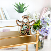 The Forged Flare® Mother's Angels® Doctor/Nurse Ornament, a 3.5" angel figurine with white wings and a pink heart, is displayed on a gold shelf amid potted plants, books, and purple flowers for a cozy decorative touch.