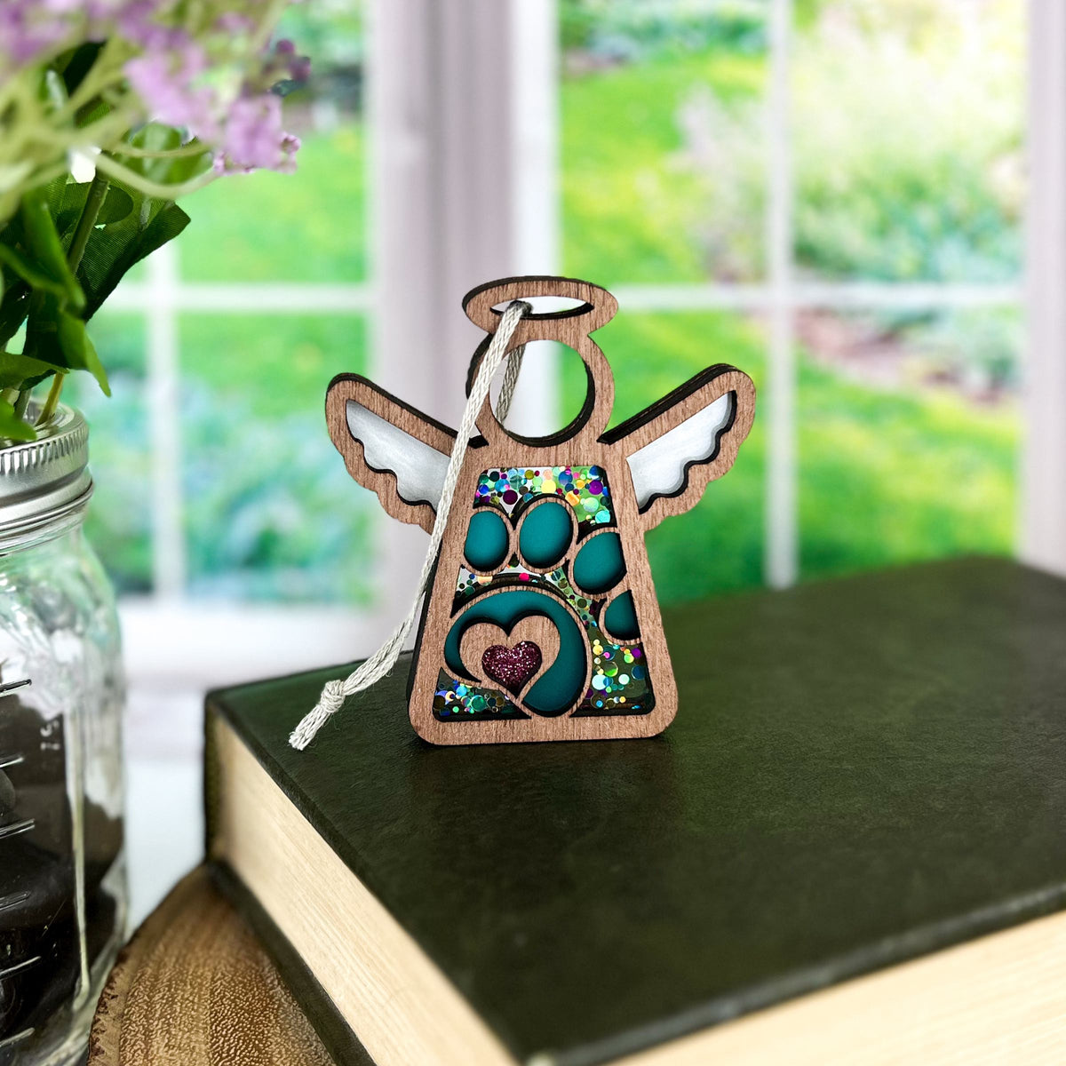 The Forged Flare® Paw Print Ornament with Imperfections—a 3.5" (H) Mother's Angels® angel figurine—rests atop a closed book, making a heartfelt pet memorial gift. A jar of flowers sits by the window overlooking the garden.