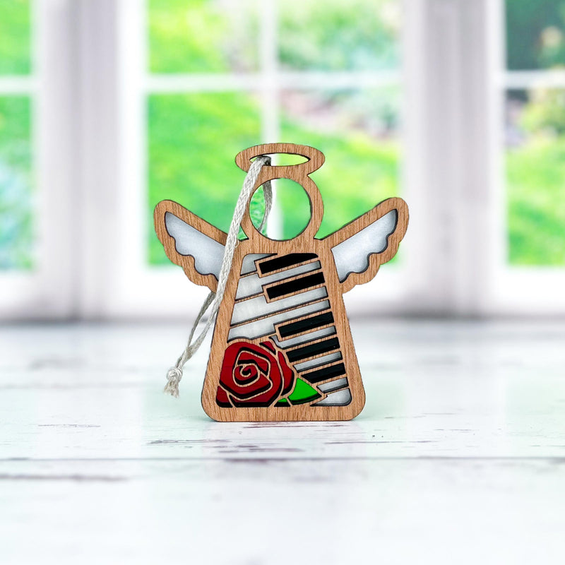 The Forged Flare® Mother's Angels® Piano & Rose Ornament with Imperfections is a 3.5" handcrafted angel figurine featuring cut-out wings, black and white piano keys, and a red rose, displayed on a white surface with a blurred window in the background.
