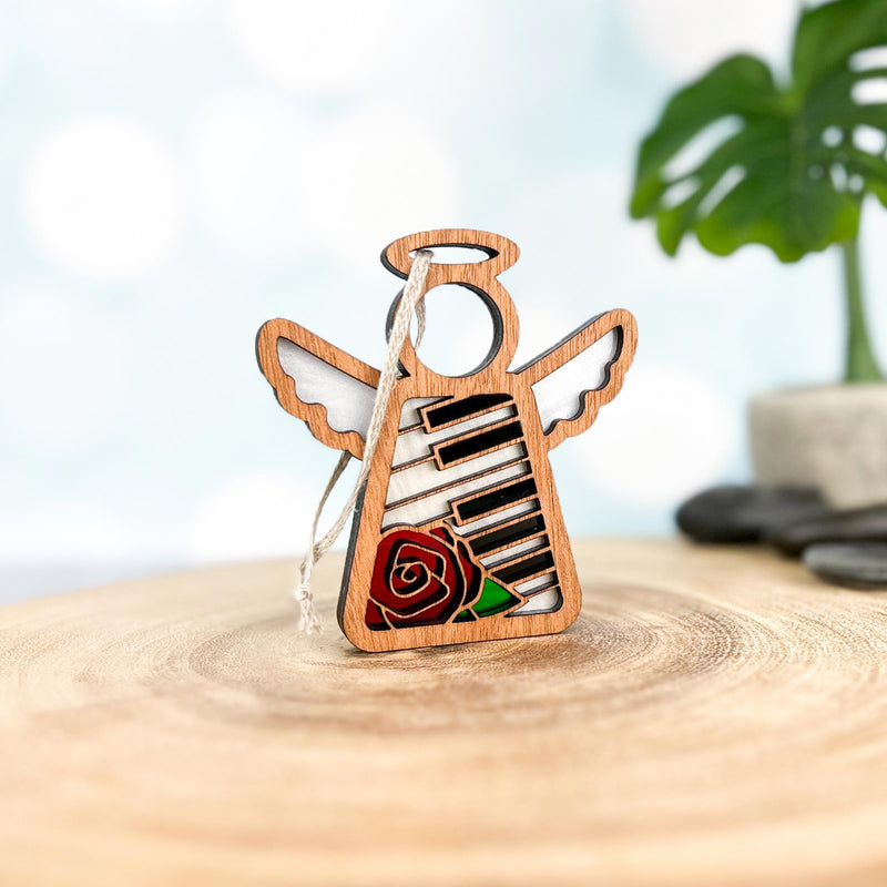 The Forged Flare® Piano & Rose Ornament with Imperfections is a 3.5" handcrafted Mother's Angels® figurine featuring piano keys and a red rose, white hanging string attached. Perfect collectible for music lovers. Green plant blurred in the background.