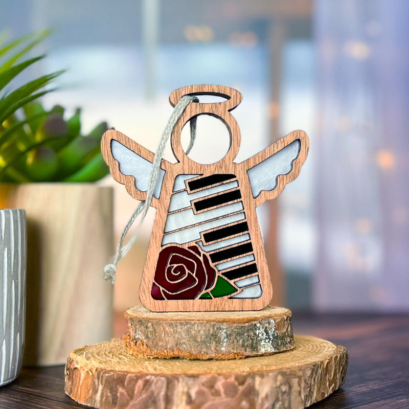 The Forged Flare® Mother's Angels® Piano & Rose Ornament with Imperfections is a 3.5" handcrafted wood angel featuring stained glass-style piano keys and a red rose, beautifully displayed on wood slices with a softly lit, plant-filled background.