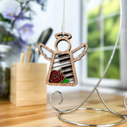 A 3.5" Forged Flare® Mother's Angels® Piano & Rose Ornament with Imperfections hangs from a silver stand on a wooden desk, with blurred flowers, a window, and art supplies in the background.