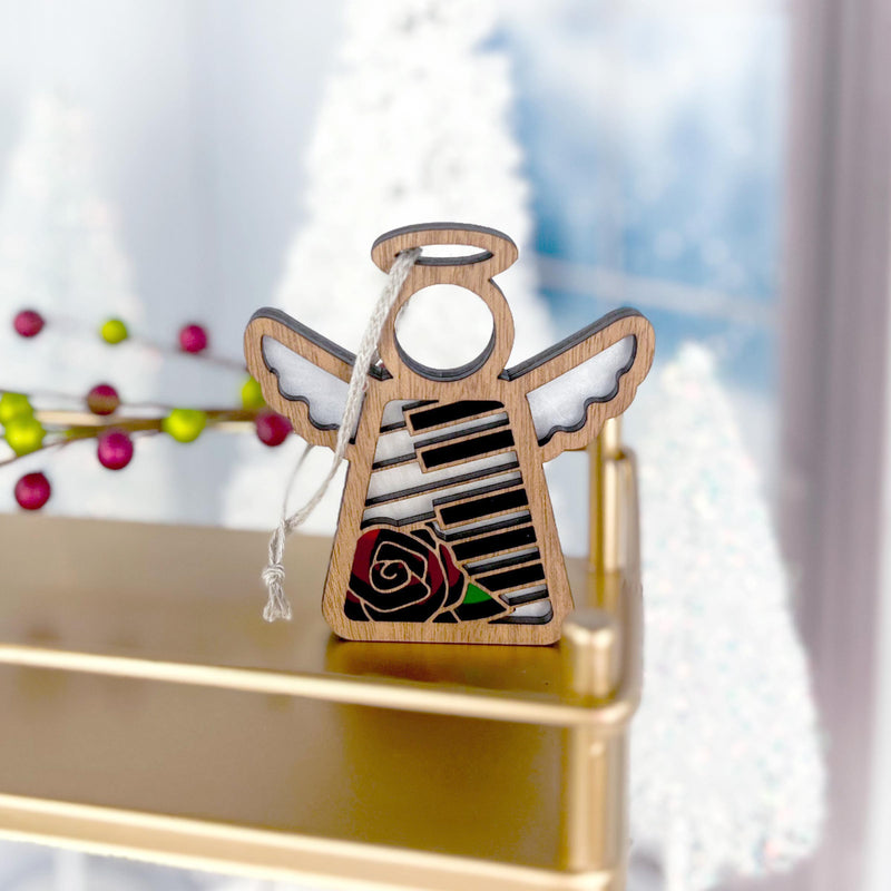 The Piano & Rose Ornament with Imperfections by Forged Flare® is a 3.5" handcrafted Mother's Angels® figurine featuring black and white piano keys and a red rose, displayed on a gold shelf with festive decor in the background.