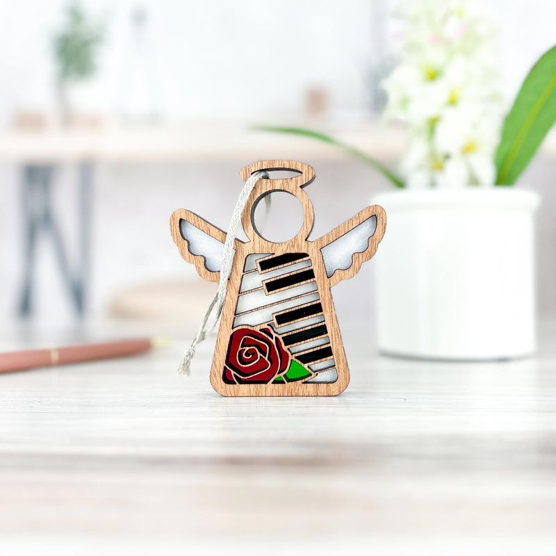The Forged Flare® Mother's Angels® Piano & Rose Ornament with Imperfections is a 3.5" handcrafted angel figurine, featuring piano keys and a red rose, displayed on a light surface with floral and pencil accents in the background.
