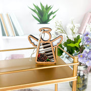 The Forged Flare® Rose & Piano Ornament—a 3.5" Mother's Angels® figurine with piano keys and red rose detail—rests on a gold shelf, ideal as music decor or a music teacher gift. Books, plants, and purple flowers appear in the background.