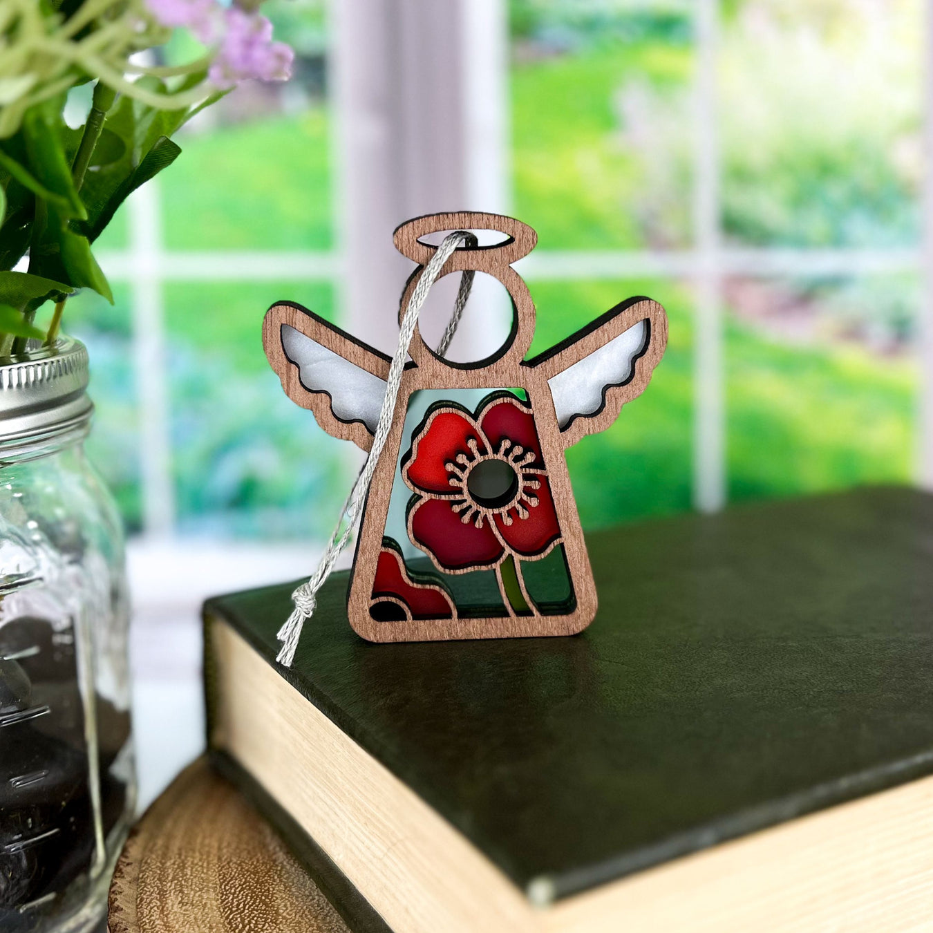 The Forged Flare® Mother's Angels® Poppy Ornament, a 3.5" stained-glass-style angel figurine with red and green details and a silver string, rests on a closed book, with flowers and a bright garden in the background. Available on Amazon.