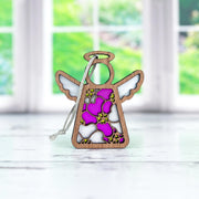 The Forged Flare® Primrose Ornament, a 3.5" Mother's Angels® figurine with white wings and a stained glass-style pink, purple, and yellow body, stands on a white surface with blurred windows and greenery behind.