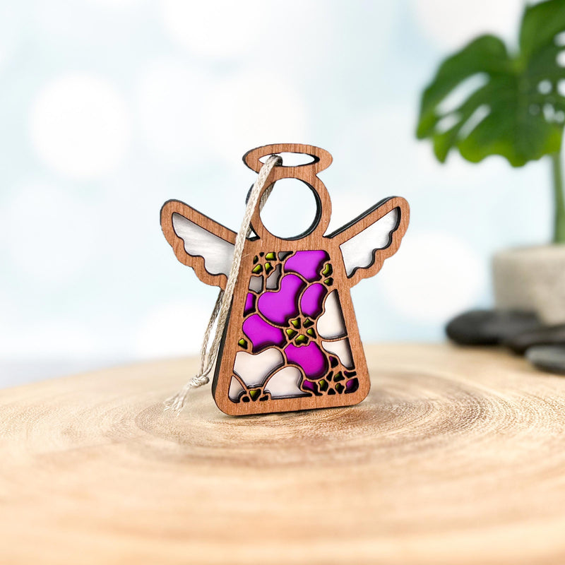 The Forged Flare® Primrose Ornament is a 3.5" wooden Mother's Angels® angel figurine with stained glass-style purple and white flowers, a pink primrose, and hanging string, displayed on wood with blurred green plant and stones in the background.