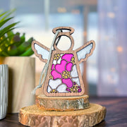 The Forged Flare® Primrose Ornament | 3.5" (H) Angel Figurine by Mother's Angels® features a stained-glass effect in pink, white, and gold, displayed on wood slices beside a green plant—a delicate tribute to the primrose flower.