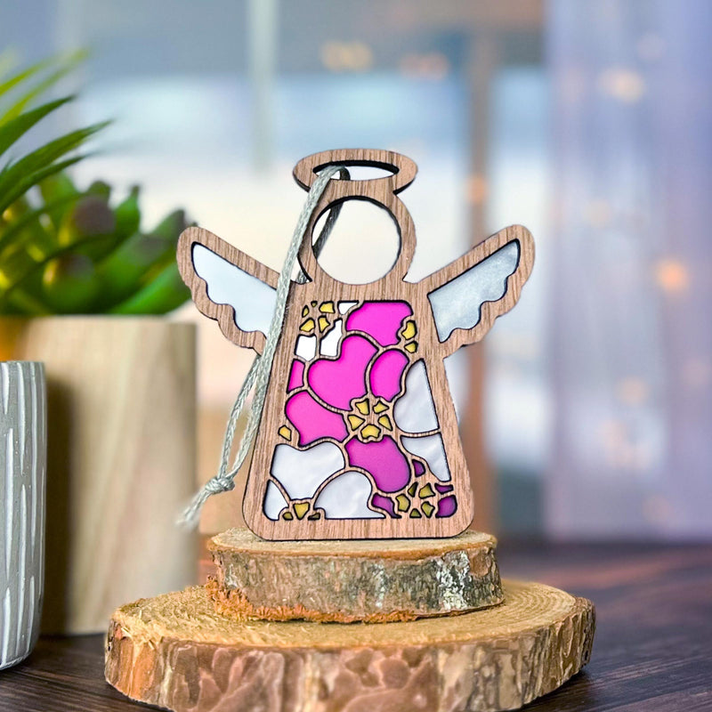 The Forged Flare® Primrose Ornament | 3.5" (H) Angel Figurine by Mother's Angels® features a stained-glass effect in pink, white, and gold, displayed on wood slices beside a green plant—a delicate tribute to the primrose flower.