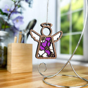 The Primrose Ornament, a 3.5" angel figurine from the Mother's Angels® collection by Forged Flare®, is adorned with intricate floral cutouts and purple accents. It hangs gracefully from a silver stand, set against a backdrop of blurred office supplies, purple flowers, and a bright window with blurred greenery. This delicate piece is an ideal birthday gift for women.
