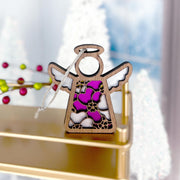 The Forged Flare® Primrose Ornament is a 3.5" Mother's Angels® figurine, featuring stained glass-style wings and body in purple, white, and pink primrose hues, hanging by a string with blurred festive decorations in the background.
