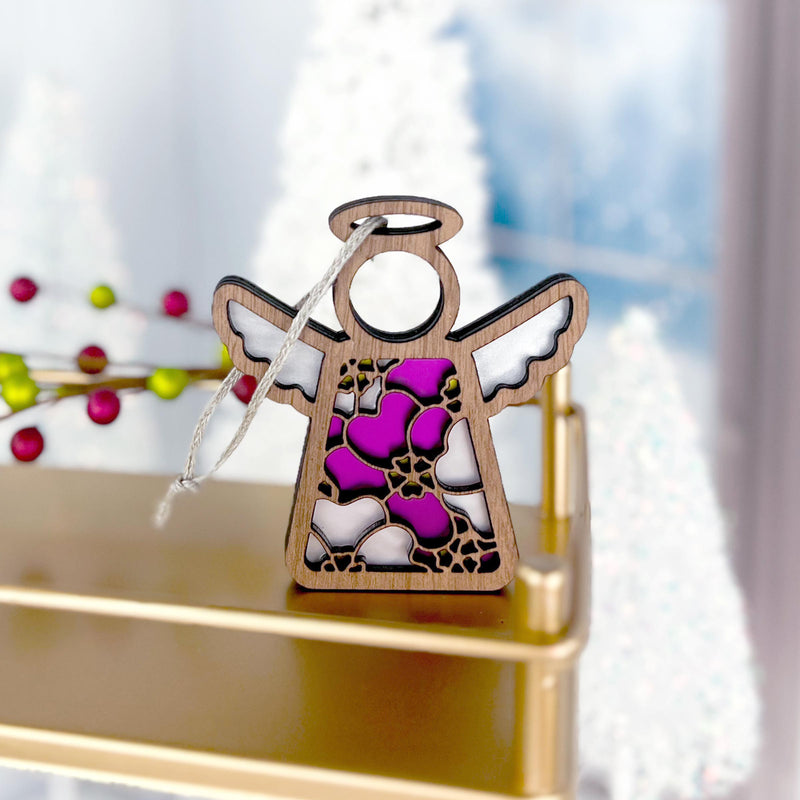 The Forged Flare® Primrose Ornament is a 3.5" Mother's Angels® figurine, featuring stained glass-style wings and body in purple, white, and pink primrose hues, hanging by a string with blurred festive decorations in the background.
