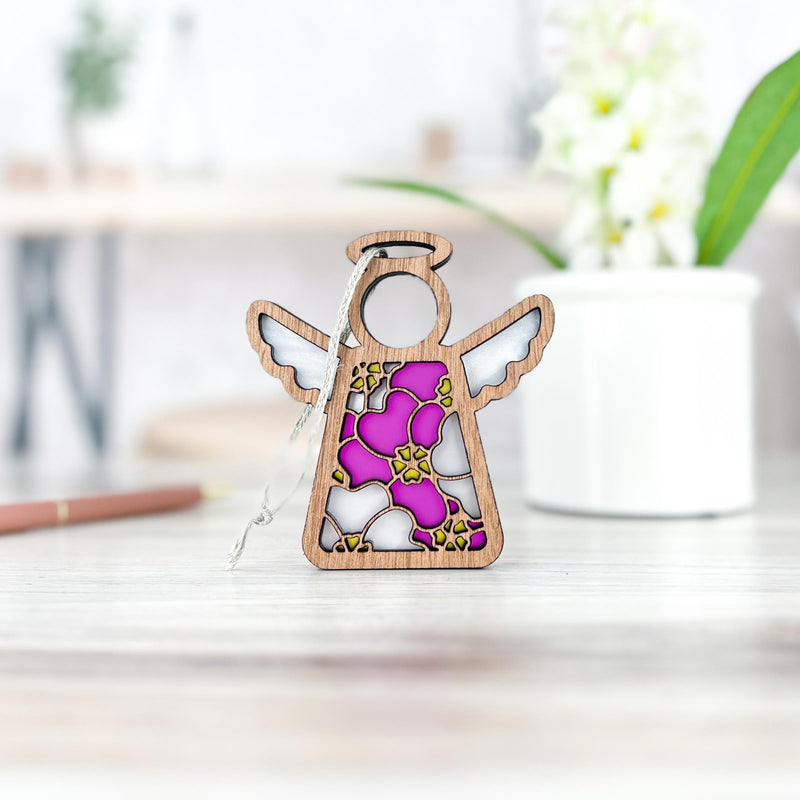 The Forged Flare® Primrose Ornament | 3.5" (H) Mother's Angels® features a wooden angel with pink, white, and gold accents and a pink primrose flower accent, standing on a table with a blurred plant pot and pen in the background.