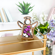 The Forged Flare® Primrose Ornament | 3.5" (H) Angel Figurine from Mother's Angels® features pink, white, and purple stained glass on a gold shelf, with books, greenery, and pink primroses in the background for a charming touch.