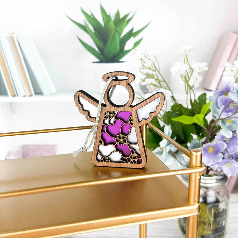 The Forged Flare® Primrose Ornament | 3.5" (H) Angel Figurine from Mother's Angels® features pink, white, and purple stained glass on a gold shelf, with books, greenery, and pink primroses in the background for a charming touch.