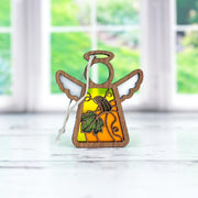 The Forged Flare® Festive Pumpkin Ornament, a 3.5" Mother's Angels® figurine with white wings, an orange body, and green leaf detail, is perfect Thanksgiving decor and sits on a light surface with greenery blurred in the background.