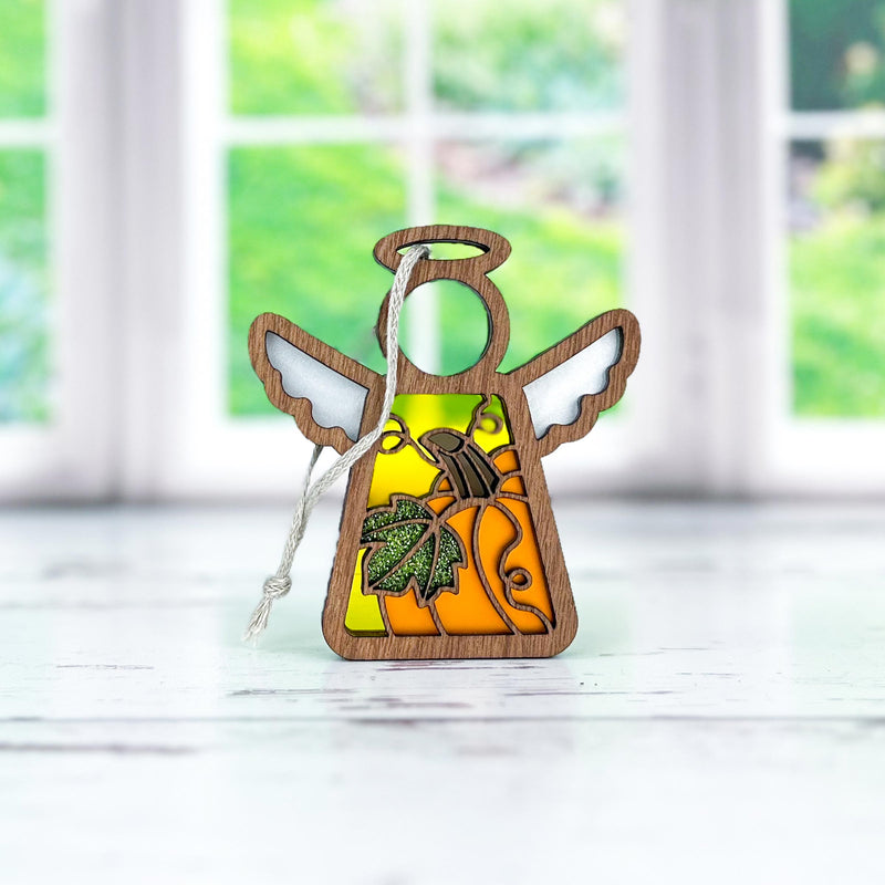 The Forged Flare® Festive Pumpkin Ornament, a 3.5" Mother's Angels® figurine with white wings, an orange body, and green leaf detail, is perfect Thanksgiving decor and sits on a light surface with greenery blurred in the background.