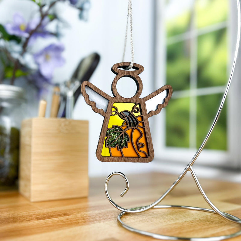 The Forged Flare® Mother's Angels® Festive Pumpkin Ornament, a 3.5" wooden angel figurine with a green leaf design and yellow-orange background, hangs on a metal stand by a window, bringing Thanksgiving charm to the decor.