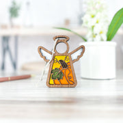 The Festive Pumpkin Ornament by Forged Flare® is a 3.5" Mother's Angels® figurine with wooden wings and a yellow-orange stained-glass body with leaf accents—ideal for fall or Thanksgiving décor. Shown on a light wooden surface with a blurred backdrop.
