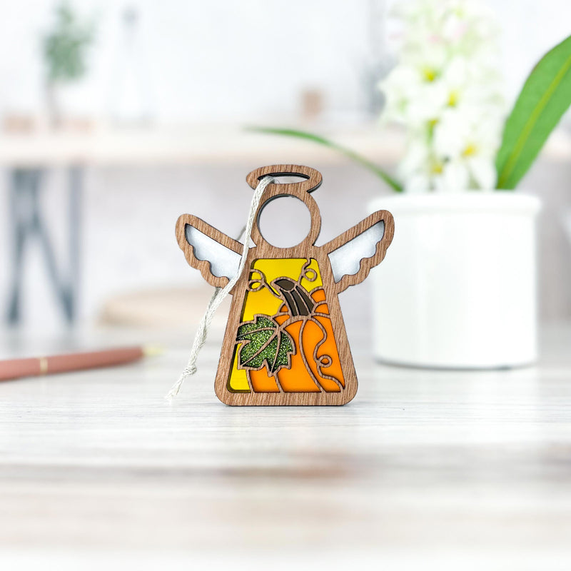 The Festive Pumpkin Ornament by Forged Flare® is a 3.5" Mother's Angels® figurine with wooden wings and a yellow-orange stained-glass body with leaf accents—ideal for fall or Thanksgiving décor. Shown on a light wooden surface with a blurred backdrop.