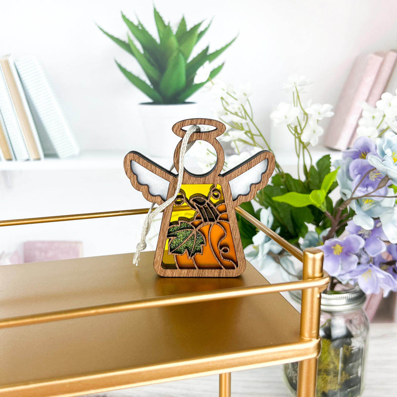 A Festive Pumpkin Ornament from Forged Flare®, showcasing a 3.5" angel figurine from the Mother's Angels® collection, adorned with intricate designs and a rope loop, sits on a gold-colored shelf. The angel features colorful autumn leaves and a pumpkin motif, making it perfect for Thanksgiving decor or as a Christmas tree ornament. Softly blurred books and white and purple flowers in a jar create an elegant background.