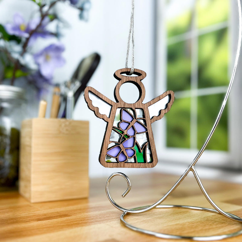 The Playful Purple Butterflies Ornament with Imperfections, a 3.5" Mother's Angels® angel figurine by Forged Flare®, featuring purple and green details, hangs from a silver stand on a desk with a blurred floral background.