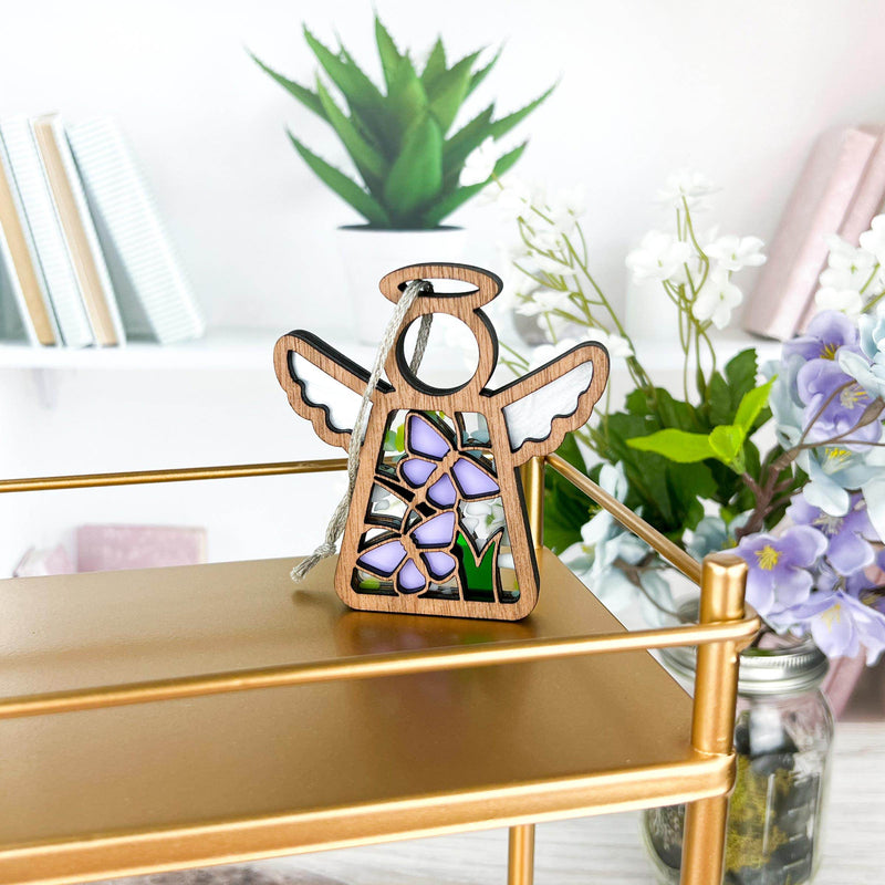 The Forged Flare® Mother's Angels® Playful Purple Butterflies Ornament (3.5"H) features purple and green stained-glass-style details and sits on a gold shelf with books, a plant, and pastel flowers in the background. Minor imperfections may be present.