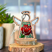 The Forged Flare® Mother's Angels® Red Rose Ornament with Imperfections is a 3.5" handcrafted angel figurine, featuring white wings and a red rose, displayed on wooden slices with a green potted plant and blurred background behind it.