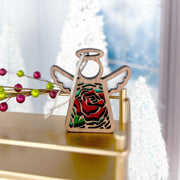 The 3.5" Red Rose Ornament with Imperfections angel figurine from Mother's Angels® by Forged Flare® stands on a gold surface, surrounded by white Christmas trees and berry decorations in a festive holiday scene.