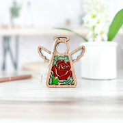 The Forged Flare® Mother's Angels® Red Rose Ornament with Imperfections is a 3.5" handcrafted wooden angel figurine featuring a red rose centerpiece and green leaves, displayed with floral accents in the background.