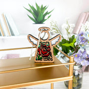 The Forged Flare® Mother's Angels® Red Rose Ornament with Imperfections, a 3.5" handcrafted angel figurine, stands on a gold shelf, surrounded by green plants, purple flowers, and books on sleek white shelves.