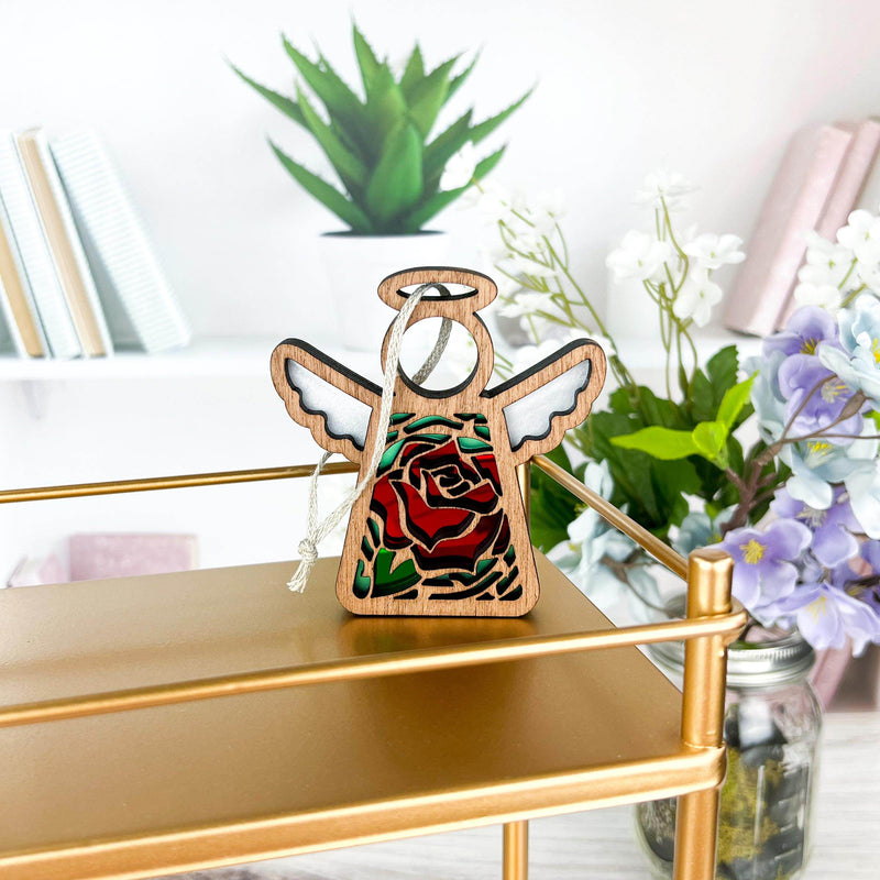The Forged Flare® Mother's Angels® Red Rose Ornament with Imperfections, a 3.5" handcrafted angel figurine, stands on a gold shelf, surrounded by green plants, purple flowers, and books on sleek white shelves.