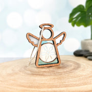 The Forged Flare® Sand Dollar Ornament with Imperfections is a 3.5” Mother's Angels® handcrafted wooden angel figurine featuring outstretched wings and a sand dollar accent—perfect for displaying against soft lights and greenery.