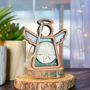 The Forged Flare® Mother's Angels® 3.5" Sand Dollar Ornament with Imperfections is a handcrafted wooden angel figurine featuring wings and a sand dollar centerpiece, shown on stacked wood slices with blurred potted plant and string lights in the background.
