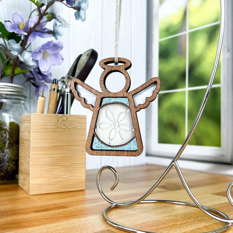 The Forged Flare® Mother’s Angels® Sand Dollar Ornament with Imperfections is a 3.5"H handcrafted angel figurine, displayed on a metal stand and wooden base, with purple flowers and a window enhancing its inviting charm.