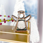 The Forged Flare® Mother's Angels® Sand Dollar Ornament with Imperfections, a 3.5" angel figurine, rests on a gold shelf among snowy Christmas trees near a window, its silver string and sand dollar center capturing the light.