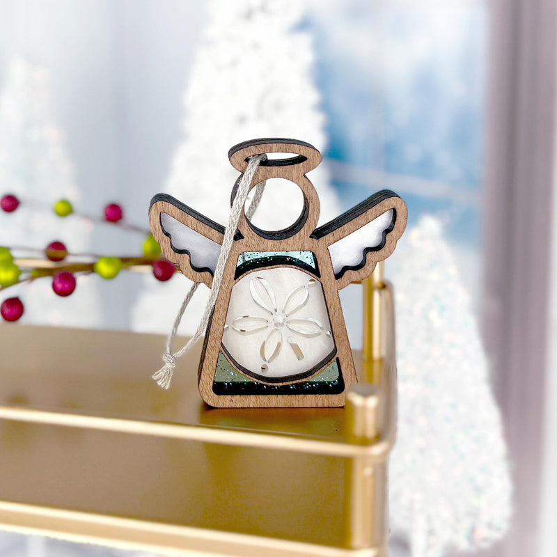 The Forged Flare® Mother's Angels® Sand Dollar Ornament with Imperfections, a 3.5" angel figurine, rests on a gold shelf among snowy Christmas trees near a window, its silver string and sand dollar center capturing the light.