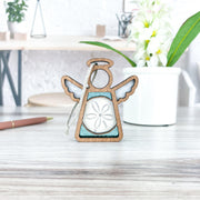 A Forged Flare® Mother's Angels® Sand Dollar Ornament with Imperfections, a 3.5" handcrafted wooden angel figurine, stands on a light desk beside a white potted plant, pen, and blurred office supplies in the background.