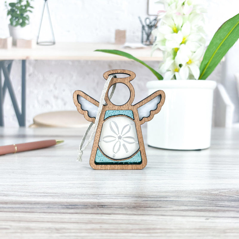 A Forged Flare® Mother's Angels® Sand Dollar Ornament with Imperfections, a 3.5" handcrafted wooden angel figurine, stands on a light desk beside a white potted plant, pen, and blurred office supplies in the background.