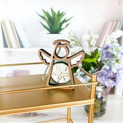 A handcrafted 3.5" Sand Dollar Ornament with white wings and a sand dollar centerpiece from Mother’s Angels® by Forged Flare® hangs on a gold shelf, with books, a potted plant, and a floral jar in the background. Ornament may have imperfections.