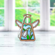 The Forged Flare® Snowdrop Ornament is a 3.5" Mother's Angels® figurine with white wings and a painted January birth flower, shown on a light surface with a blurred window and green garden in the background.