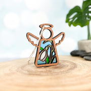 The Forged Flare® Snowdrop Ornament is a 3.5" Mother's Angels® wooden angel figurine with white wings, halo, and a stained glass-style snowdrop birth flower design, standing on wood with a blurred green plant in the background.