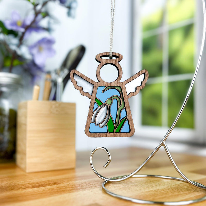 The Snowdrop Ornament from the Mother's Angels® collection by Forged Flare® is a 3.5" angel figurine with wooden edges, featuring white snowdrop flowers on a blue background. It hangs from a stand, set against a backdrop of a window with green foliage and a desk adorned with art supplies and a small wooden box, making it an ideal birthday gift for women.