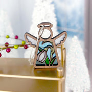 The Forged Flare® Mother's Angels® Snowdrop Ornament is a 3.5" wooden angel figurine with white wings and a snowdrop flower, displayed on gold with blurred white trees and red berries in the snowy background.